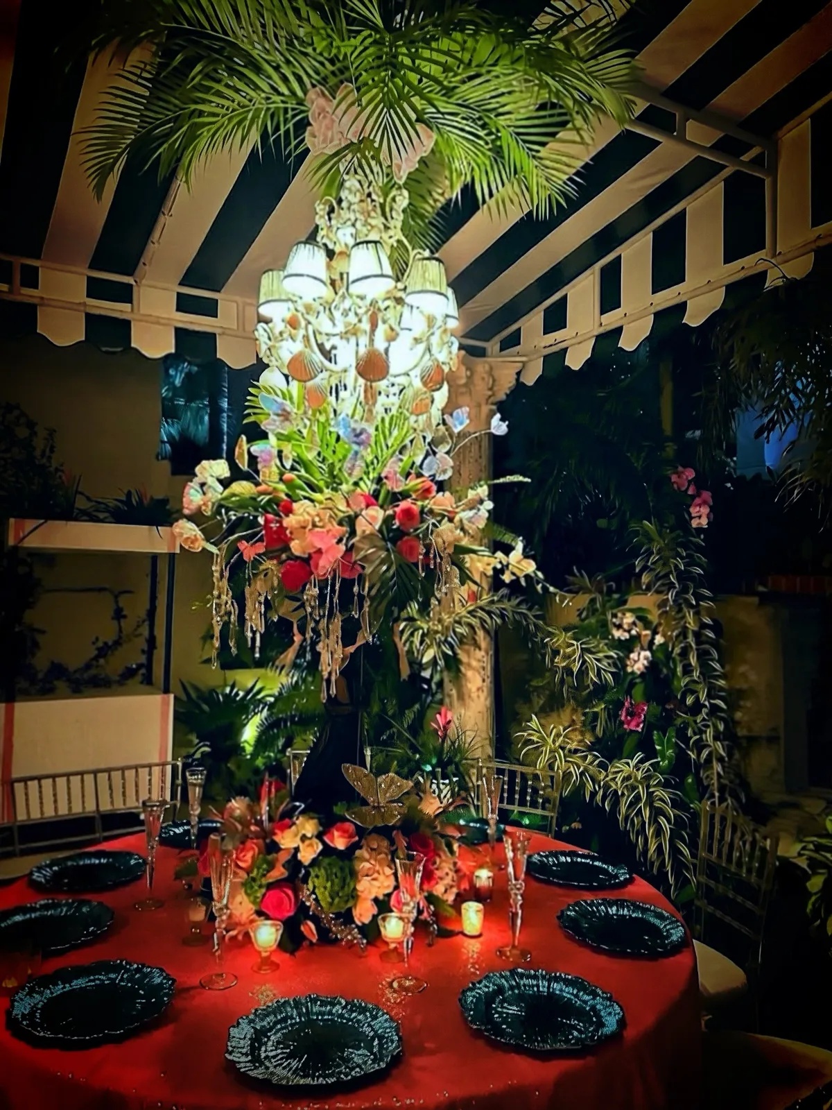 A dramatic after-dark tablescape with a chandelier, tropical foliage, florals, candles, and black place settings.