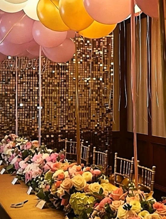 A long event table with floral arrangements, gold sequin backdrop, and blush and gold balloons.