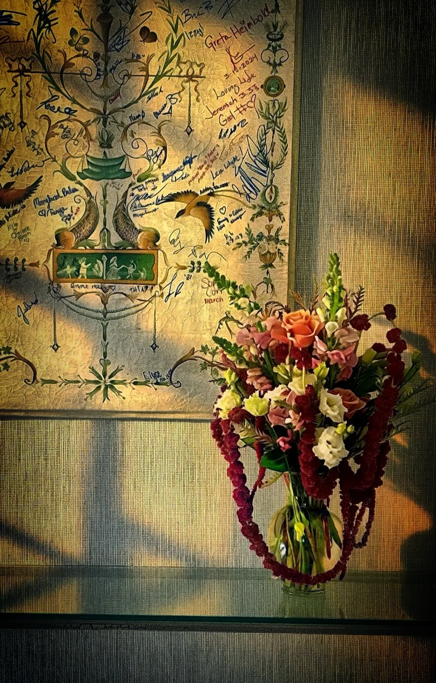 A warm hospitality floral arrangement with peach, burgundy, and cream flowers against an art-filled wall.